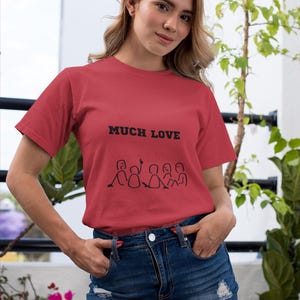 May include: A red t-shirt with the words "MUCH LOVE" printed in black, and a line drawing of people. The model is wearing a denim skirt. The background includes greenery and a white wall.