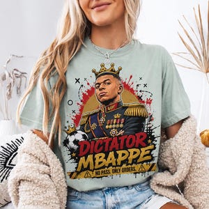 May include: A pale green t-shirt with a graphic of a man in a crown and military uniform, with the words "DICTATOR MBAPPE" in red and yellow. A football is also in the graphic.
