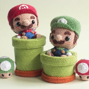 May include: Two crocheted figures, one wearing a red hat and one wearing a green hat, each emerging from a green cylindrical pipe. Two small crocheted mushrooms, one green and one red, are positioned on the sides of the two pipe structures.