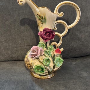 Puede incluir: Un jarrón decorativo de cerámica con cuerpo crema, ribete dorado y asa curvada. El jarrón está adornado con rosas rosas y burdeos tridimensionales, hojas verdes y una pequeña flor roja.