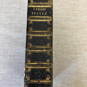 Könnte beinhalten: Ein schwarzer, ledergebundener Buchrücken mit goldfarbenen Prägedetails und horizontalen Bändern. Der Text "COMMON PRAYER" ist in goldenen Großbuchstaben auf dem oberen Teil des Buchrückens gedruckt, der mit dekorativen Schnörkelmustern versehen ist.