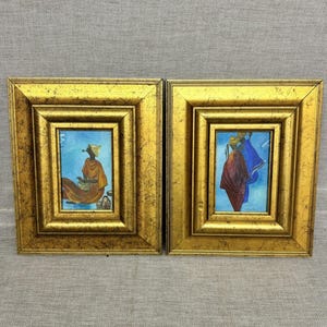 Può includere: Due dipinti rettangolari in cornici dorate ornate e anticate. Il dipinto a sinistra raffigura una figura con un indumento arancione su sfondo blu. Quello a destra mostra due figure con vesti rosse e blu su uno sfondo blu.