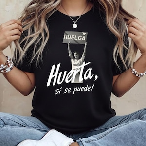 May include: Black t-shirt featuring a graphic of a woman holding a sign that says "HUELGA" above her head. Below the image is the text "Huerta, sí se puede!" in white script. The shirt is worn by a person wearing blue jeans.