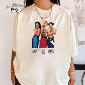May include: An ivory-coloured t-shirt featuring a graphic print of three individuals in colourful outfits, including blue and red trousers, with three distinct signatures printed below the figures. The text COMFORT COLOR Ivory is printed in the top left corner.