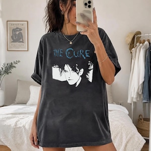 May include: A charcoal gray short-sleeve t-shirt featuring a graphic print of a person with dark hair and the text "THE CURE" in blue lettering. The shirt has a crew neckline and a relaxed, oversized fit with a washed-out, vintage-style fabric finish.