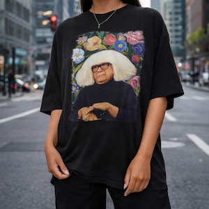 May include: Black t-shirt featuring a portrait of a man with white hair and glasses, surrounded by colorful flowers. The man is wearing a black turtleneck. The shirt is a casual, oversized fit.