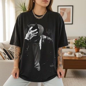 May include: Black t-shirt featuring a black and white image of a person wearing a hat and jacket, with a signature on the right side. The shirt is a dark color and has a relaxed fit.