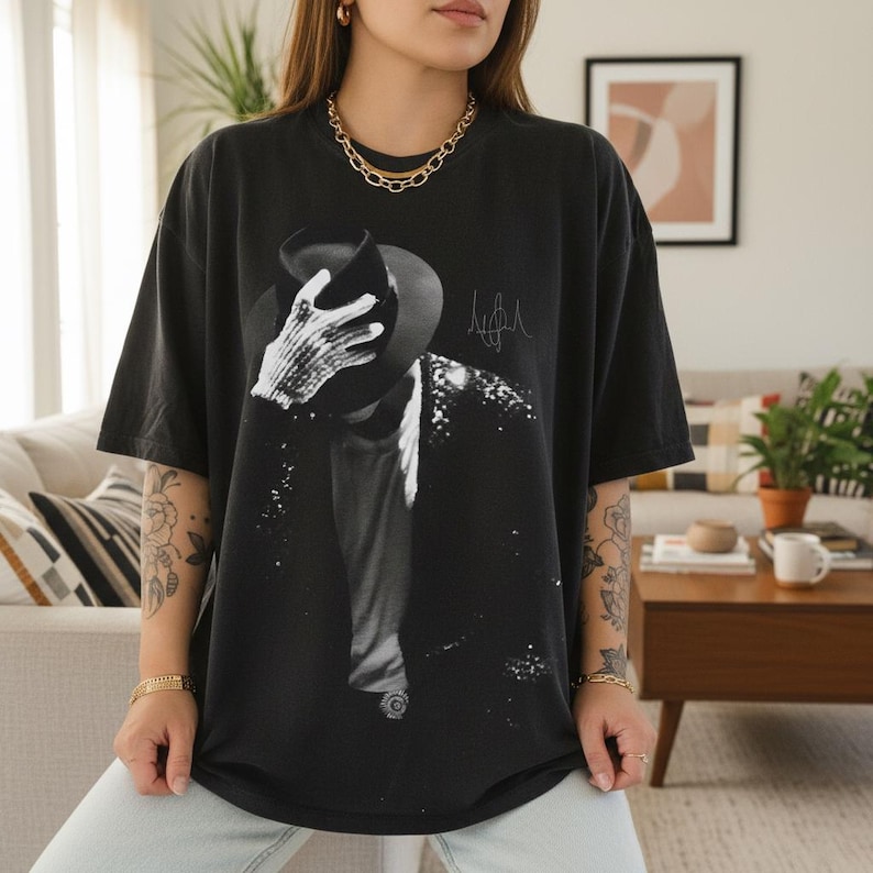 May include: Black t-shirt featuring a black and white image of a person wearing a hat and jacket, with a signature on the right side. The shirt is a dark color and has a relaxed fit.