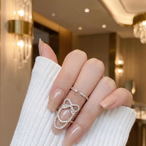 Pode incluir: Uma mão usando dois anéis de prata com pedras preciosas transparentes. Um anel é uma banda delicada, e o outro é um anel de unha com um design de laço. As unhas estão pintadas com uma cor neutra.