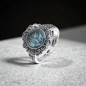 Pode incluir: Um anel prateado com um design ornamentado e vazado. O anel apresenta uma pedra redonda azul-esverdeada no centro, rodeada por uma cravação de filigrana detalhada. A banda é ajustável, com um design dividido. O anel está sobre um fundo neutro.