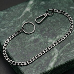Può includere: Una catena in metallo color argento con un moschettone e un portachiavi. La catena è composta da maglie ad incastro ed è esposta su una superficie di marmo verde.
