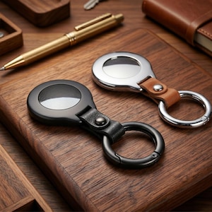Pode incluir: Dois chaveiros com design circular, um preto e outro prateado, cada um preso a uma tira de couro e um anel de metal. Uma caneta dourada e um caderno de couro também são visíveis na superfície de madeira.