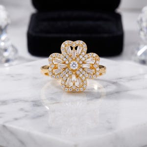 Pode incluir: Um anel dourado com um design de trevo de quatro folhas. O trevo é incrustado com pequenas pedras redondas e baguete transparentes. O anel está sobre uma superfície de mármore branco, com uma caixa de joias preta ao fundo.