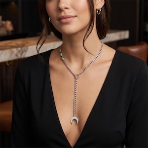 Pode incluir: Colar prateado com um pendente de lua crescente. O colar tem um design de corrente de elos com uma queda em forma de Y. A modelo usa um top preto e brincos de argola prateados.