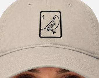 May include: A beige baseball cap featuring a black and white bird illustration inside a black rectangle, with the number 1 in the upper left corner. The cap is made of a soft material.
