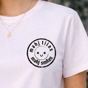 May include: White t-shirt with a circular black graphic that reads "mahj tiles mahj smiles" around a smiling face. The shirt is plain and has a crew neck.