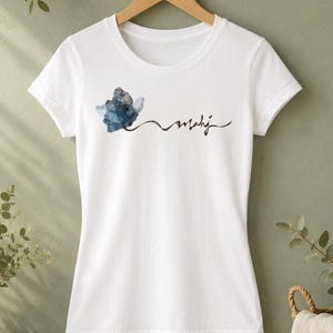 May include: White t-shirt with a crew neck, featuring a watercolor-style blue and gray floral design and the word "mahij" in a handwritten font. The shirt is hanging on a wooden hanger against a green background.