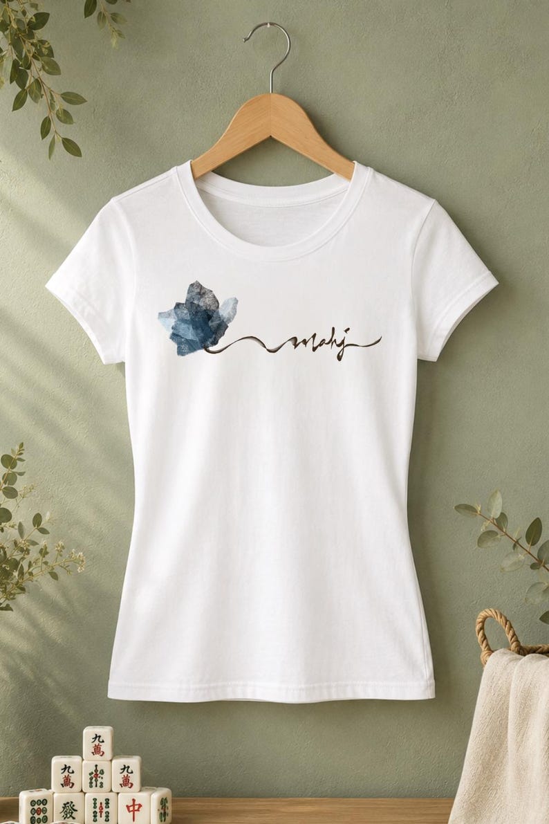 May include: White t-shirt with a crew neck, featuring a watercolor-style blue and gray floral design and the word "mahij" in a handwritten font. The shirt is hanging on a wooden hanger against a green background.