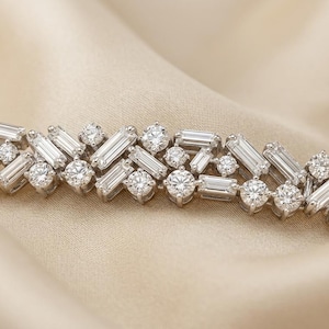 Könnte beinhalten: Ein silberfarbenes Armband mit einem sich wiederholenden Muster aus Baguette-Schliff-Steinen in Chevron-Anordnung, durchsetzt mit runden Brillantschliff-Steinen. Die Metallfassung ist poliert und reflektierend und bildet ein durchgehendes, bewegliches Band.