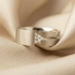 Könnte beinhalten: Ein silberfarbener Ring mit gebürstetem, mattem Finish und einem geteilten Ringschienen-Design. Ein einzelner, klarer, dreieckig geschliffener Edelstein ist zwischen den beiden offenen Enden des Rings gefasst und bildet einen geometrischen Mittelpunkt.