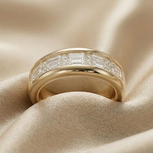 Könnte beinhalten: Ein Goldring mit einem zentralen Kanal, der mit einer Reihe rechteckiger Baguette-geschliffener klarer Steine besetzt ist, die sich mit kleinen runden klaren Steinen abwechseln. Das polierte Goldmetall hat ein glattes, abgerundetes Profil.
