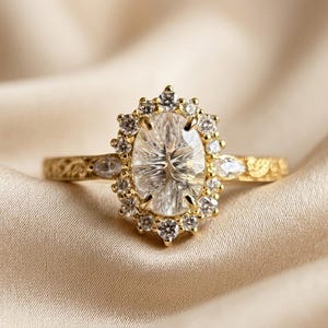 Könnte beinhalten: Ein Goldring mit einem großen oval geschliffenen Mittelstein, der von einem Kranz aus kleinen, runden, klaren Steinen umgeben ist. Die Ringschiene besteht aus Gelbgold mit einem filigranen Blumenmuster und zwei marquise-geschliffenen Steinen.