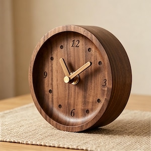 May include: A circular wooden clock in a dark brown hue. The clock face has gold-coloured hands and hour markers. The numbers 3, 6, 9, and 12 are visible. The clock is placed on a light brown surface.