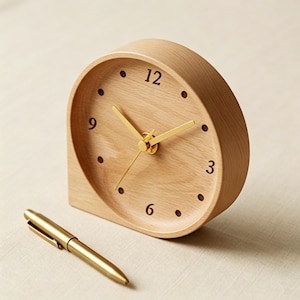 May include: A light-colored wooden desk clock with a rounded top and a flat bottom. The clock face has black dots for hour markers and gold-colored hands. A gold pen rests on the surface next to the clock.