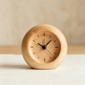 May include: A small, round wooden clock with a light brown finish. The clock face is also light brown with black hands and simple markings for the hours. The clock has a minimalist design and would be a nice addition to a desk or nightstand.