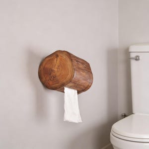 Puede incluir: Un portarrollos de papel higiénico de madera rústica, hecho de una sección de tronco de árbol. El soporte está montado en una pared gris claro, con papel higiénico blanco que sale de la abertura. La veta natural de la madera y el grano circular son visibles.