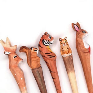 Puede incluir: Colección de cinco figuras de animales de madera talladas a mano. Las figuras incluyen un ciervo, una ardilla, un tigre, un gato y un conejo. Cada figura está pintada con colores naturales y detalles. Fondo blanco.