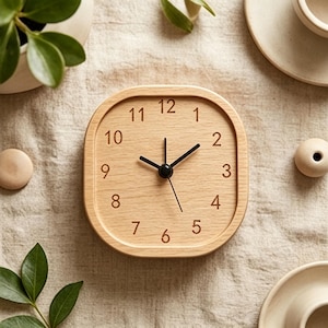 May include: A square wooden clock with rounded corners. The clock face is light-colored wood with black numerals and hands. The clock is surrounded by neutral-toned decor, including a small plant and wooden accents.