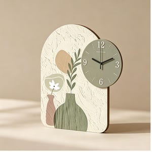May include: A decorative wall clock featuring a textured off-white background with painted botanical elements and a round, sage green clock face. The clock has a wooden frame and a minimalist design, suitable for home decor.