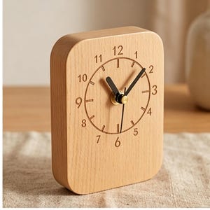 May include: A rectangular wooden desk clock with a light brown finish. The clock face features black numerals and hands, with a gold-colored center. The clock is set on a light-colored surface.