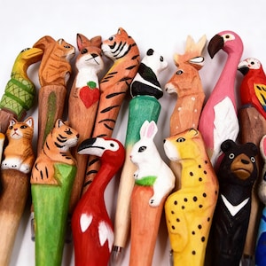 Könnte beinhalten: Eine Sammlung handgeschnitzter Holzstifte mit Tieraufsätzen. Die Stifte zeigen eine Vielzahl von Tieren, darunter Tiger, Panda, Flamingo und Bär, mit leuchtenden Farben und detaillierter Handwerkskunst. Jeder Stift ist einzigartig und sticht hervor.