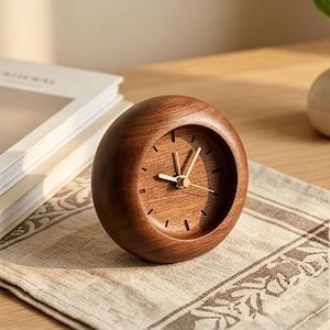 May include: A small, round wooden clock with a dark brown finish. The clock face is visible, featuring gold-colored hands and simple hour markers. The clock is resting on a patterned beige surface, suggesting a decorative or functional item.