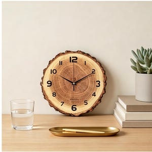 May include: A round wooden clock with a natural bark edge. The clock face features black numerals and hands, with a visible wood grain pattern. The clock is on a wooden surface with a glass of water, a gold tray, and a succulent plant.
