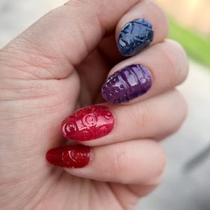 Puede incluir: Primer plano de una mano con uñas elaboradamente diseñadas. Las uñas están pintadas en varios colores, incluyendo rojo, morado, azul y verde. Cada uña presenta un diseño texturizado único, creando un efecto llamativo y artístico. Tema: belleza y moda.