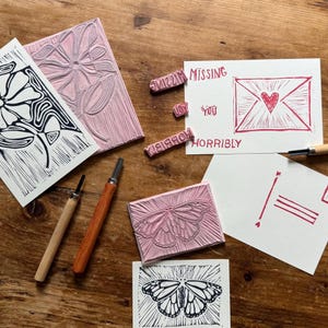 May include: Linocut printmaking supplies including two pink rubber carving blocks with flower and butterfly designs, two carving tools with wooden handles, and white cards featuring black and red ink prints of flowers, butterflies, and the text MISSING YOU HORRIBLY.