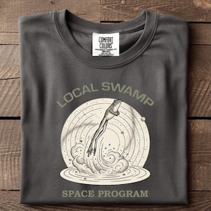 May include: Dark grey t-shirt with a circular graphic of a frog leaping from water, with the words "LOCAL SWAMP SPACE PROGRAM". The shirt is folded on a wooden surface.