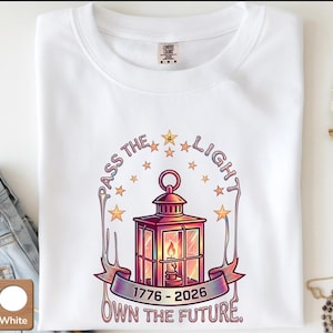 May include: White t-shirt with a graphic design featuring a lantern, stars, and the text "PASS THE LIGHT, OWN THE FUTURE" and the dates "1776-2026". The lantern is red and gold with a lit candle inside. The shirt is folded.