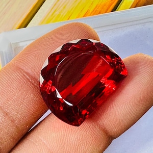 May include: A faceted, heart-shaped, deep red gemstone. The gem is held in a hand, showcasing its brilliant cut and vibrant color. The gemstone is displayed in a clear plastic container.