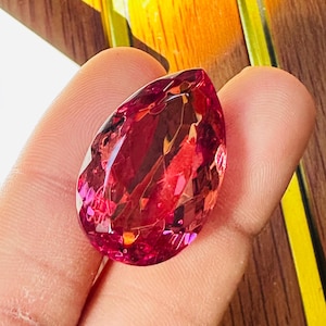 May include: A faceted, teardrop-shaped gemstone in a vivid red-pink colour. The gem's surface reflects light, displaying internal patterns and facets. The stone is held in a hand, with a wooden surface in the background.