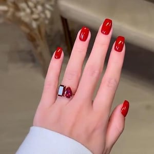 May include: A ring featuring two distinct red gemstones set on a metallic band. One stone is a rectangular baguette cut, and the other is a pear-shaped cut. The ring is worn on a finger with bright red nail polish, and the sleeve of a white garment is visible.