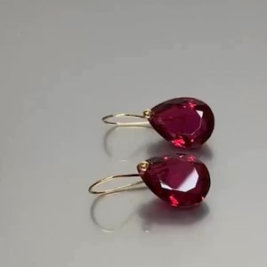 May include: A pair of teardrop-shaped earrings featuring deep red, faceted gemstones. Each earring is suspended from a thin, curved gold-tone wire hook that attaches to the top of the stone with a small, round gold-tone bead accent.