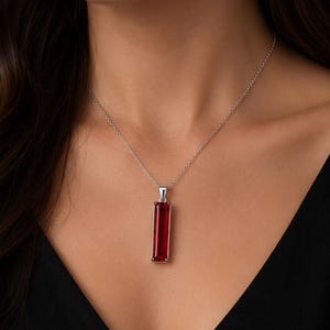 May include: A silver necklace featuring a long, rectangular, deep red gemstone pendant. The pendant is held by a simple silver bail attached to a delicate silver chain. The vibrant red stone has a smooth, polished surface and a clear, faceted appearance.