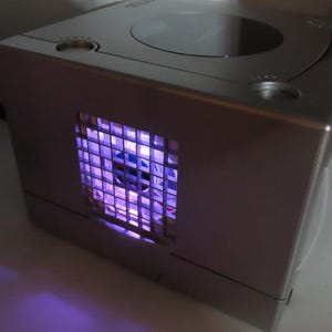 May include: Silver-colored video game console with a square shape and a circular disc tray on top. The front of the console has a grid-like design with a purple glow emanating from within. The console is sitting on a white surface.
