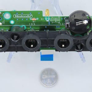 May include: A green Nintendo circuit board with black controller ports and a black component. The board has various electronic components, including resistors and a small blue connector. A silver coin is at the bottom.