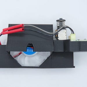 May include: A black electronic component with a fan, wires, and a spring. The fan has a blue and white label. Red wire covers are visible. The component is on a white background.