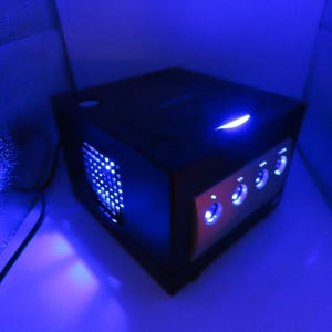 May include: A black gaming console with a blue light on top and blue lights on the front and side. The console has several circular buttons with blue lights. The overall theme is gaming.